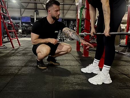 Mateusz przykucnięty przy sztangielce poprawia ustawienie podopiecznej podczas ćwiczenia